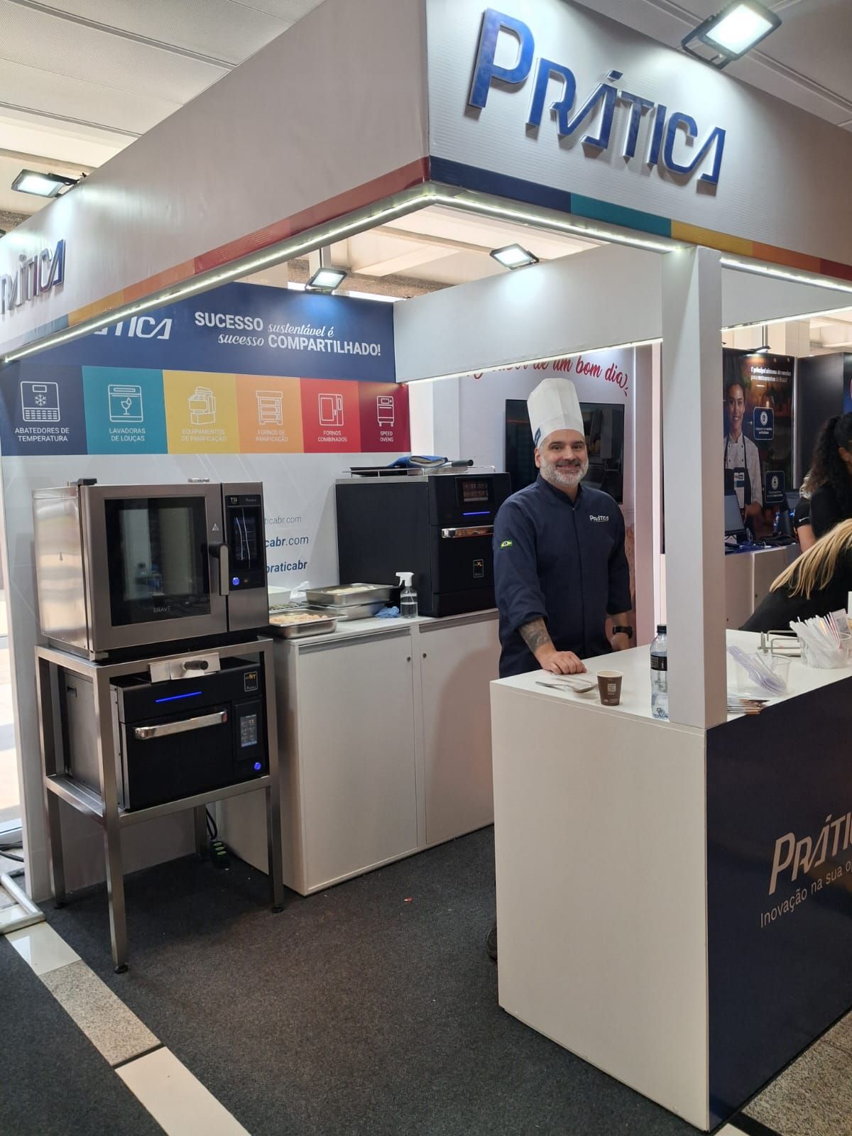 Chef no estande da Prática, exibindo fornos. Uniforme azul, chapéu branco. Interior da feira, decoração em branco e azul.