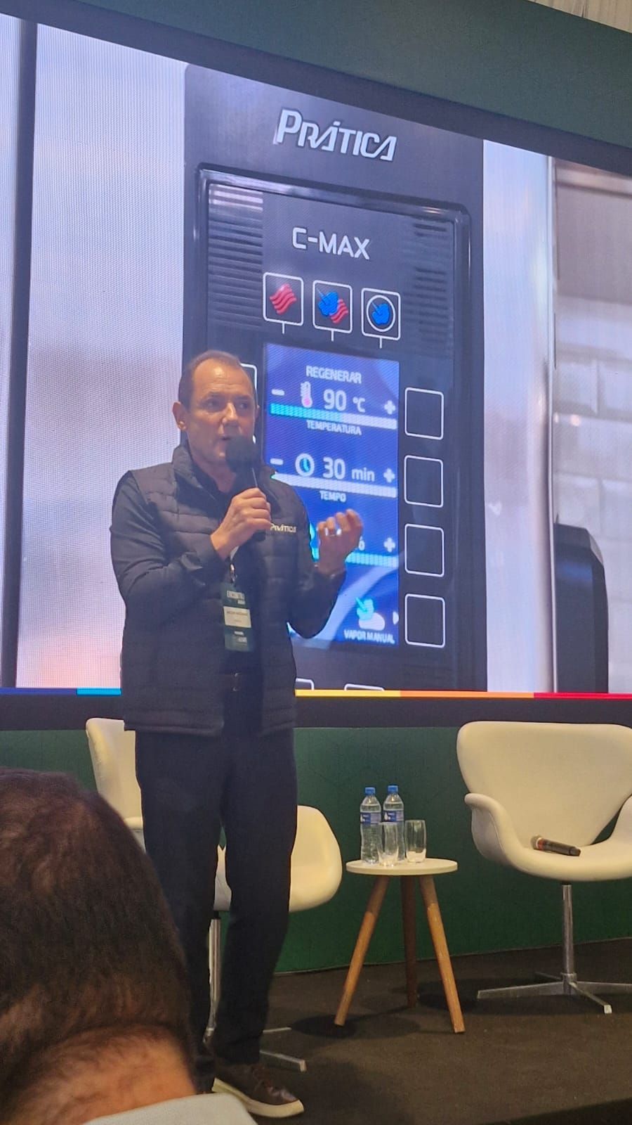 Homem falando em frente a uma vitrine, possivelmente em uma feira comercial, com uma placa de produto.