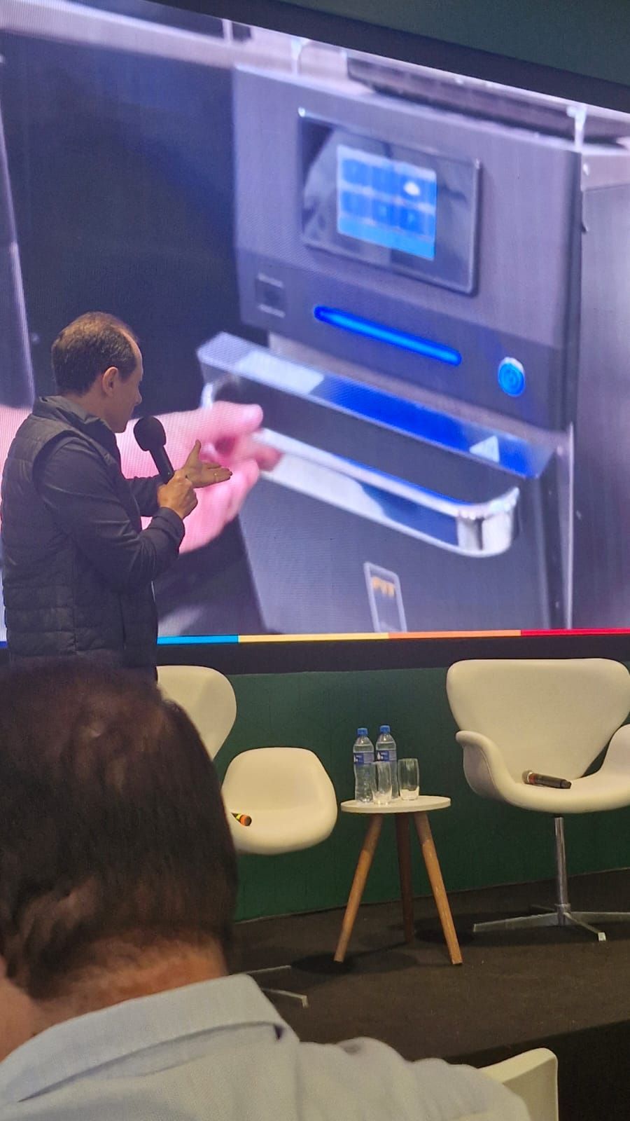 Homem fazendo uma apresentação ao lado de uma tela que mostra um dispositivo. Ele segura um produto e gesticula. Cadeiras modernas e uma pequena mesa estão em primeiro plano.