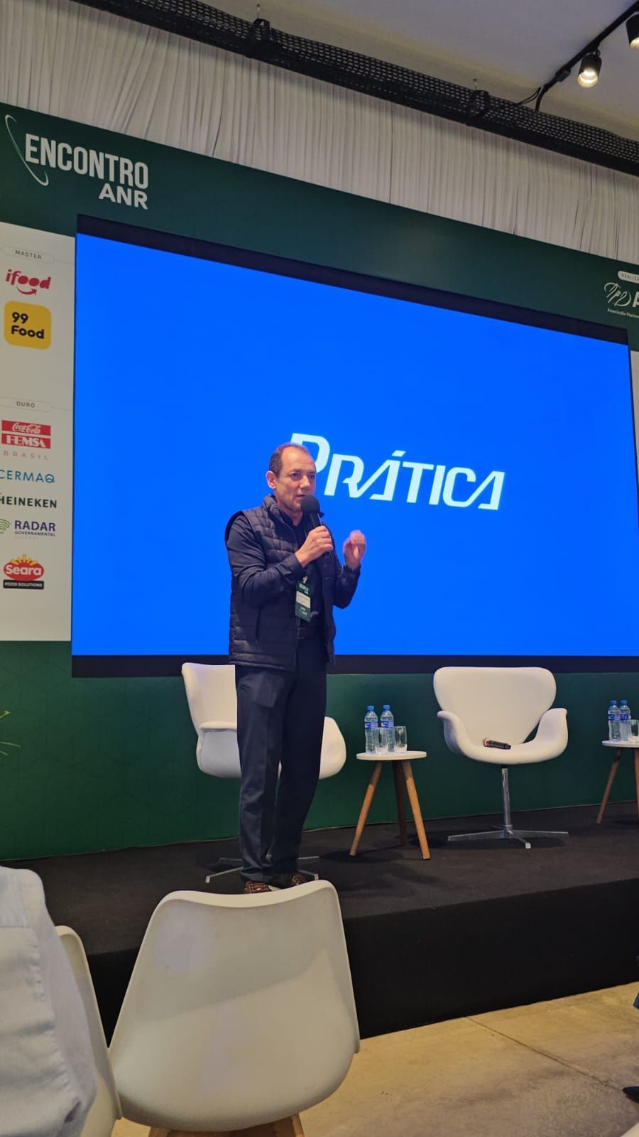 Homem falando em uma conferência, em pé sobre uma cadeira. Tela azul com a inscrição 