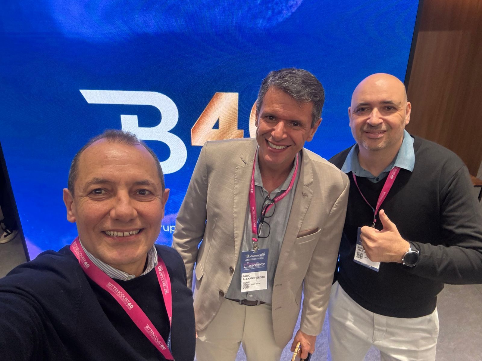 Três homens sorrindo para uma selfie em uma conferência com o logotipo 