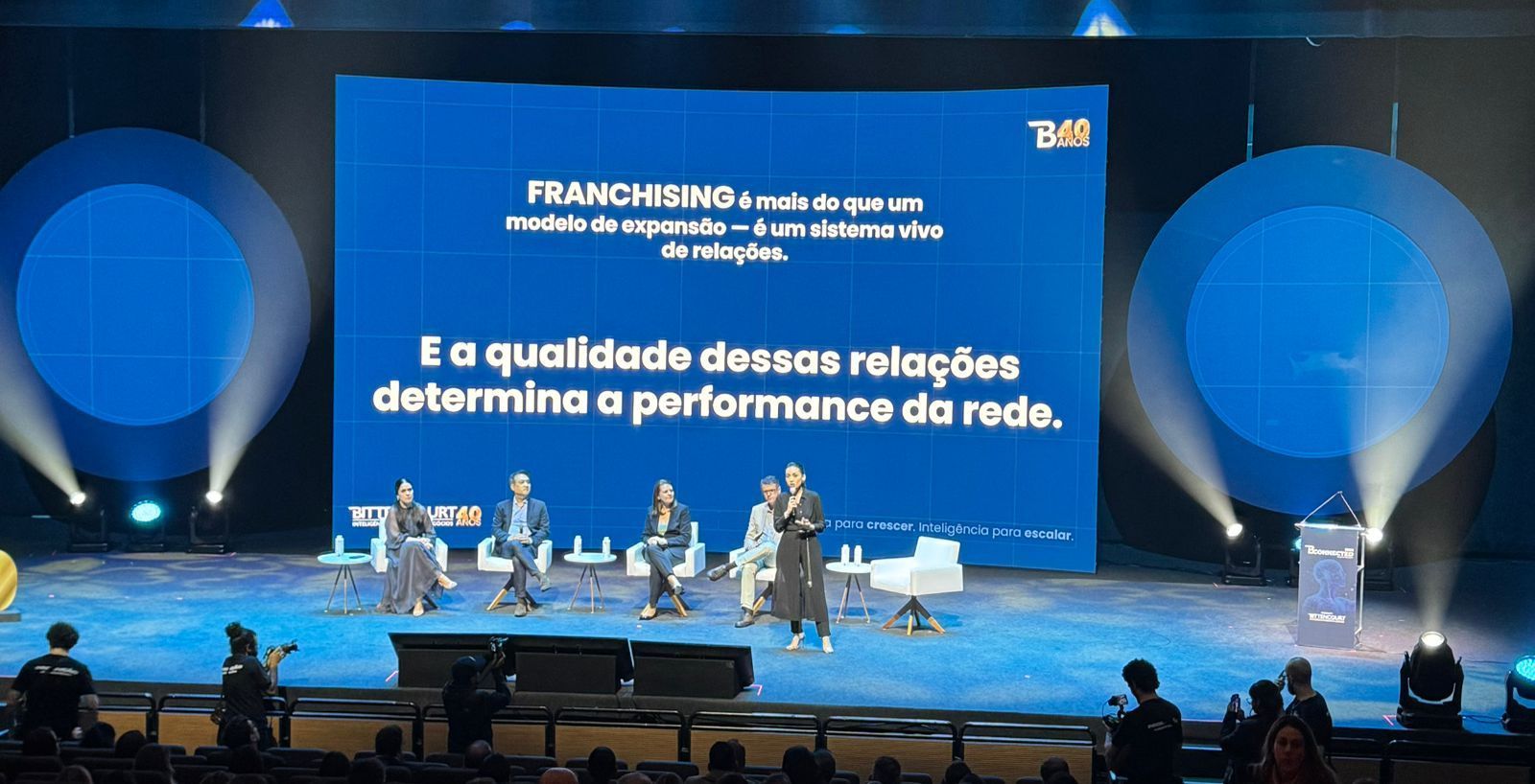Palco com palestrantes, tela azul e plateia; apresentação sobre franquias.