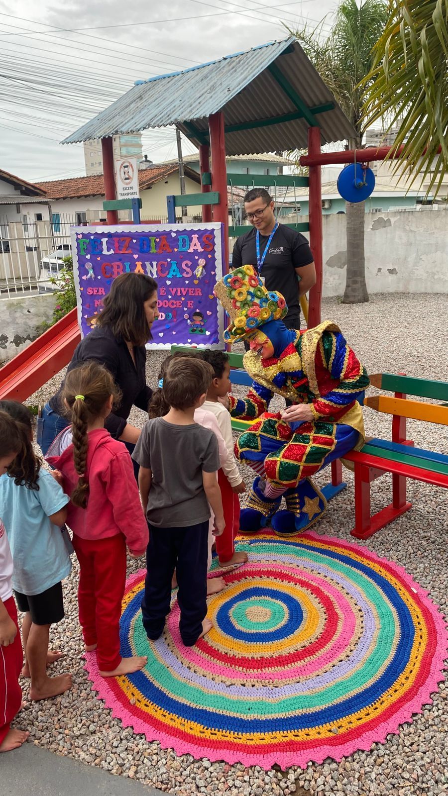 Palhaço interagindo com crianças ao ar livre, perto de um playground. Outros observam. Tapete colorido, placa, adultos.