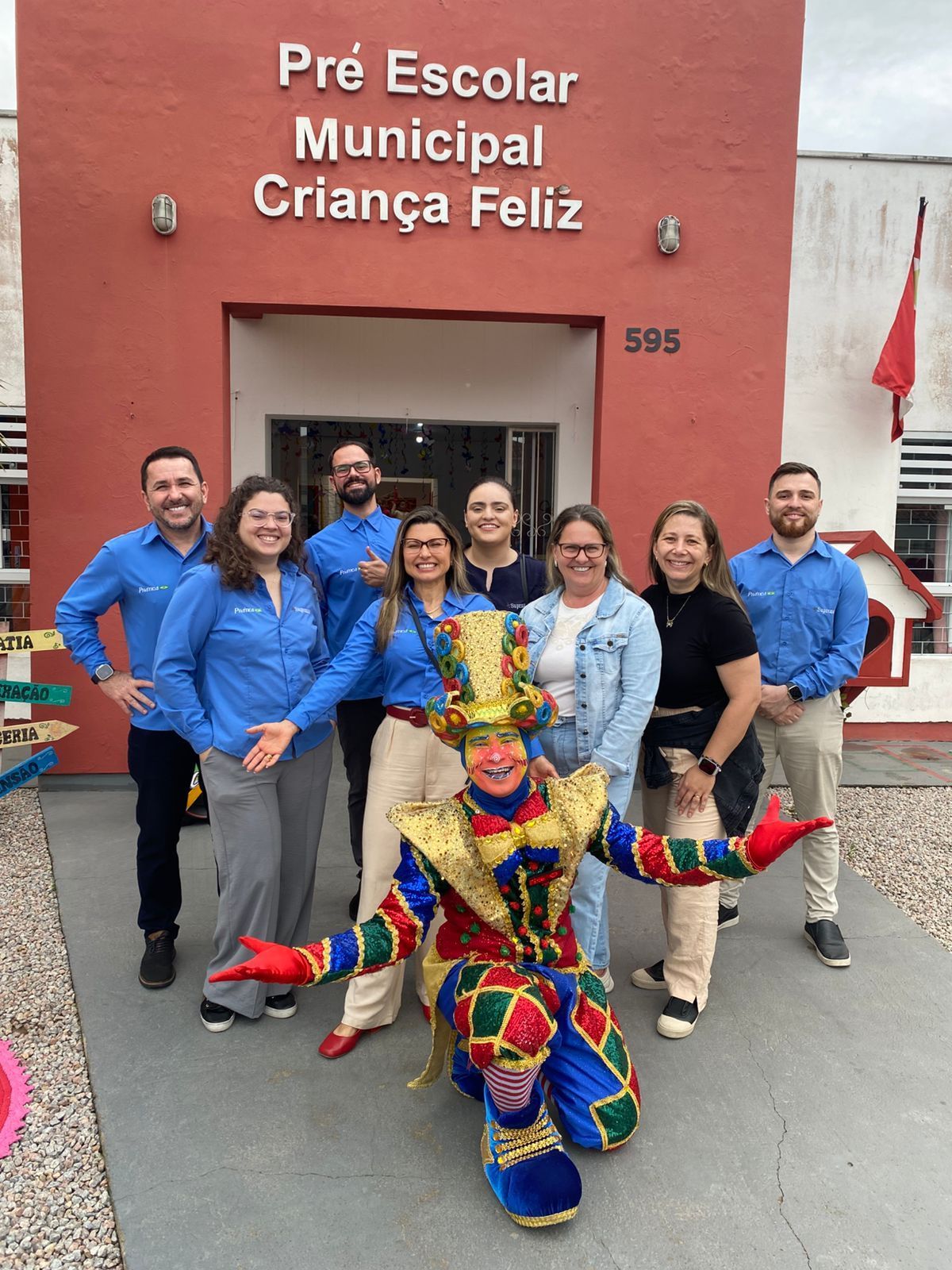 Grupo posa com palhaço em frente ao prédio vermelho de uma pré-escola.