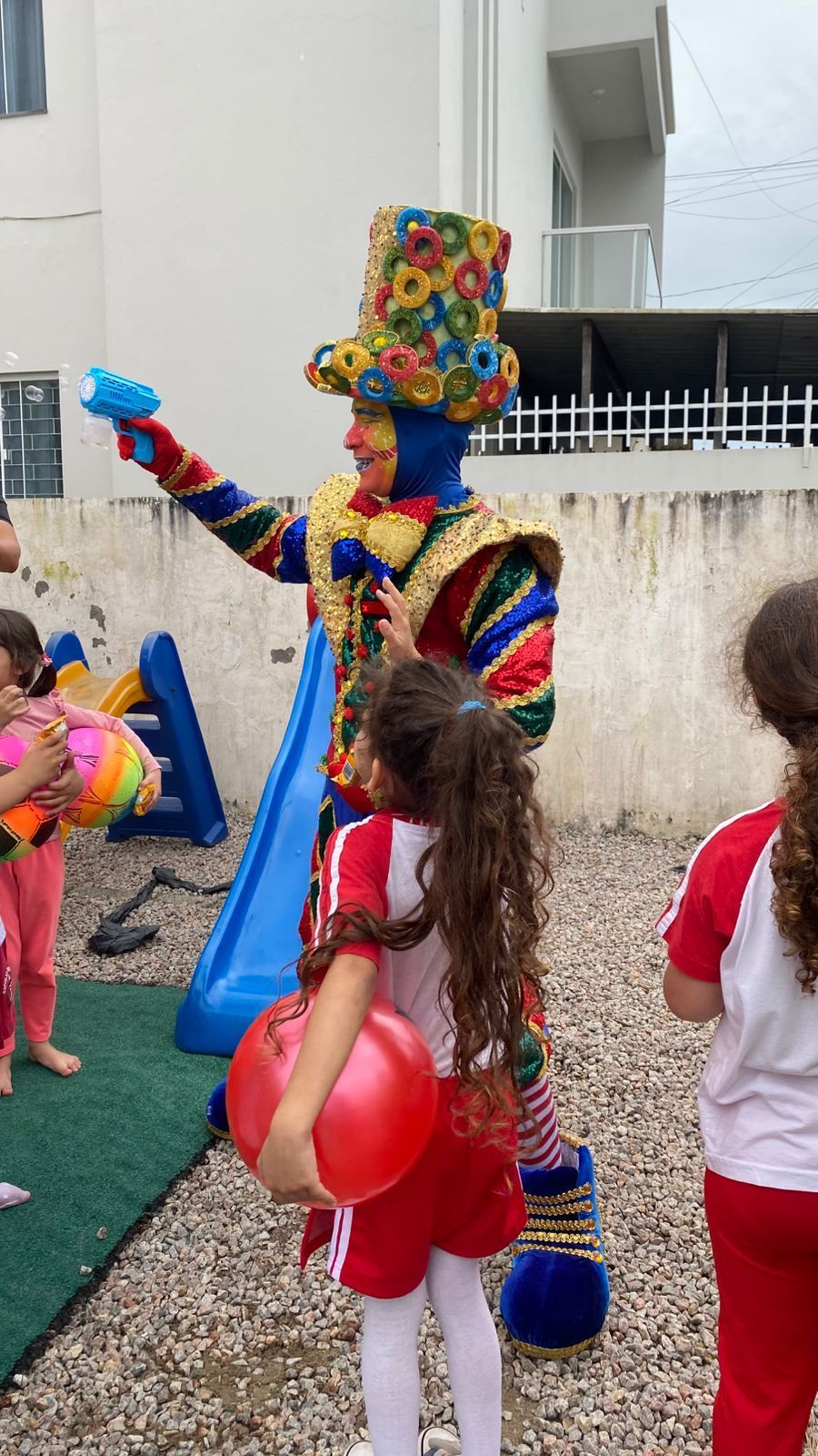Palhaço entretendo crianças do lado de fora. Palhaço usa fantasia e chapéu coloridos. Crianças seguram balões.