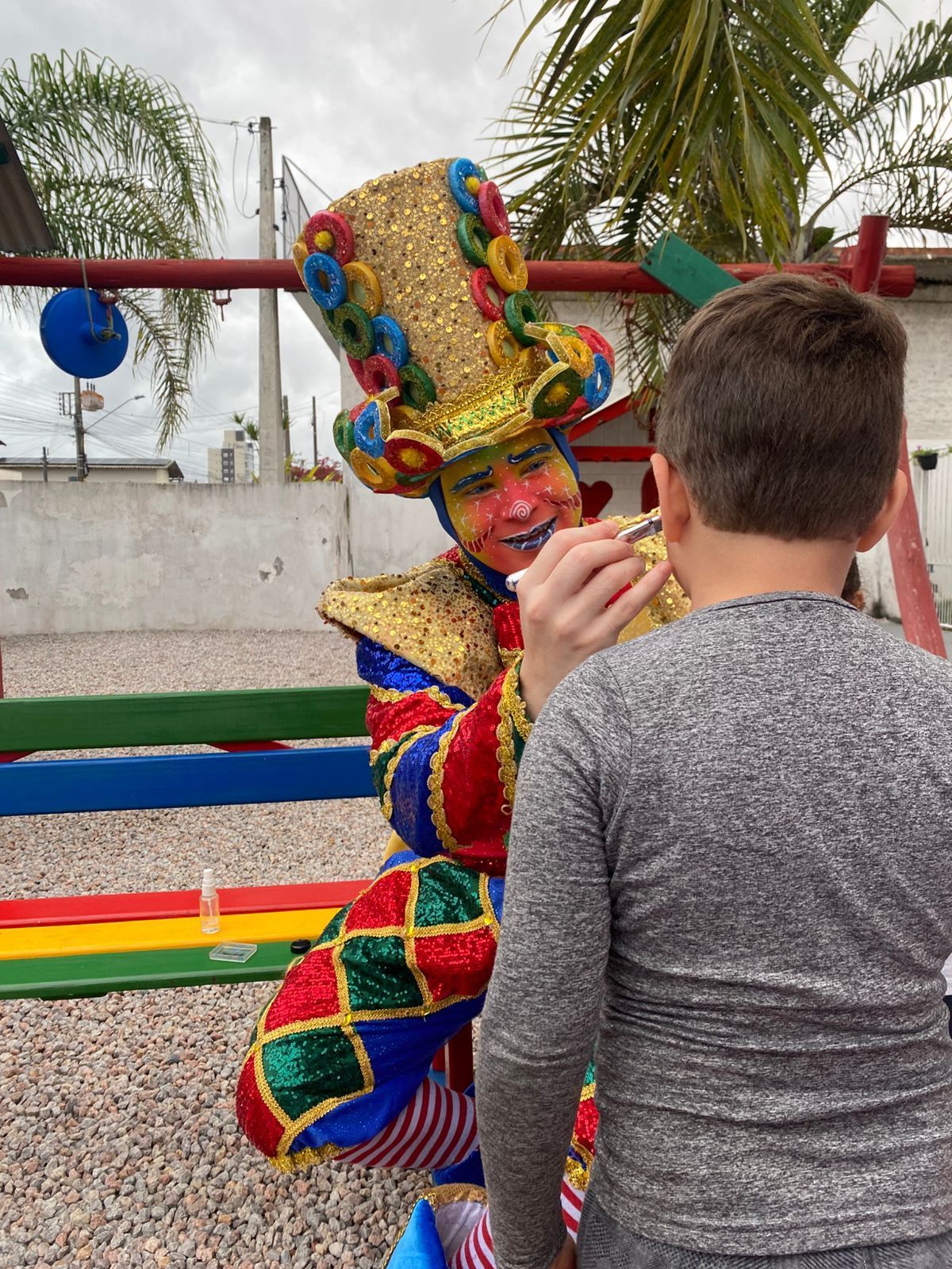 Palhaço aplicando tinta facial em um menino em um ambiente externo colorido com um playground.