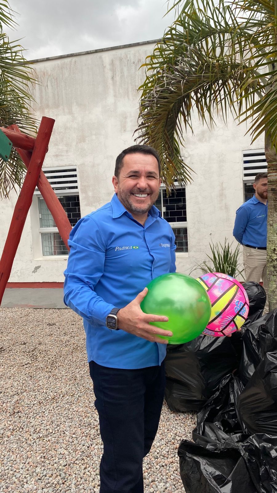Homem de camisa azul sorri, segurando uma bola verde, ao lado de outras bolas e sacos de lixo. Ao ar livre.