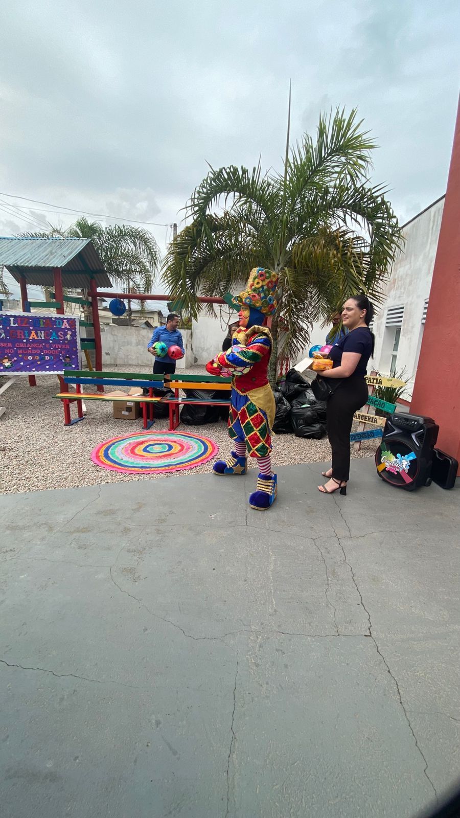 Palhaço fazendo malabarismo com bolas ao ar livre, com uma mulher e uma pessoa atrás. O palhaço é colorido, com vermelho e amarelo, e faz malabarismo com bolas vermelhas.