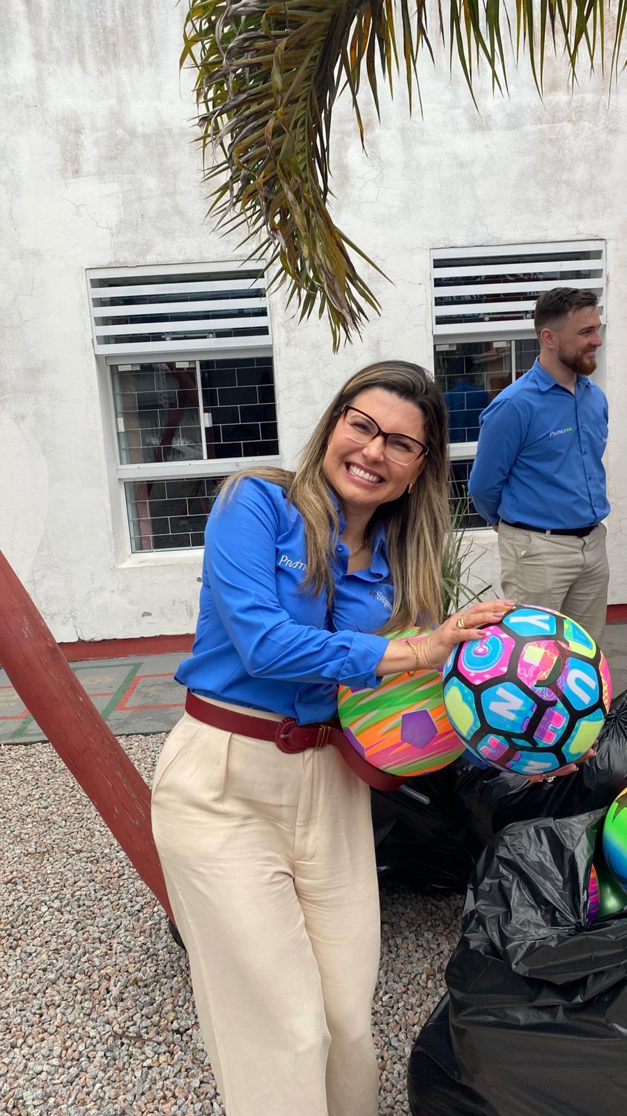 Uma mulher de camisa azul e óculos sorri, segurando uma bola colorida; outro homem está por perto.