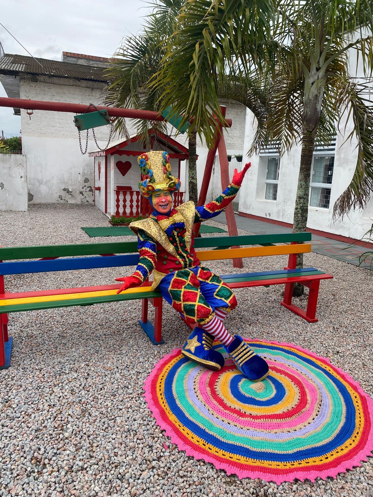 Uma pessoa com uma fantasia colorida de arlequim está sentada em um banco de arco-íris em um quintal, perto de um tapete.
