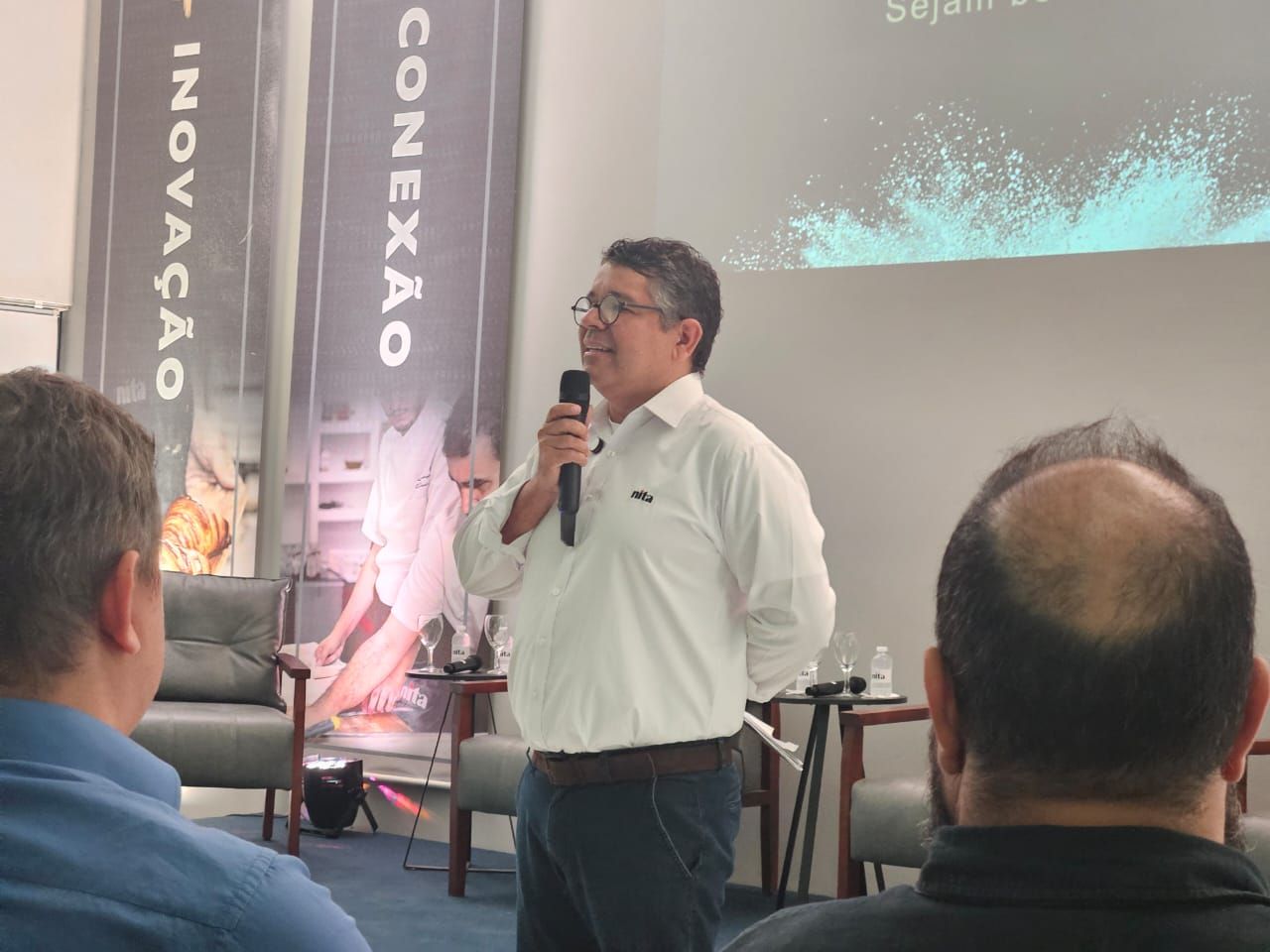 Homem falando ao microfone em evento. Banner ao fundo com