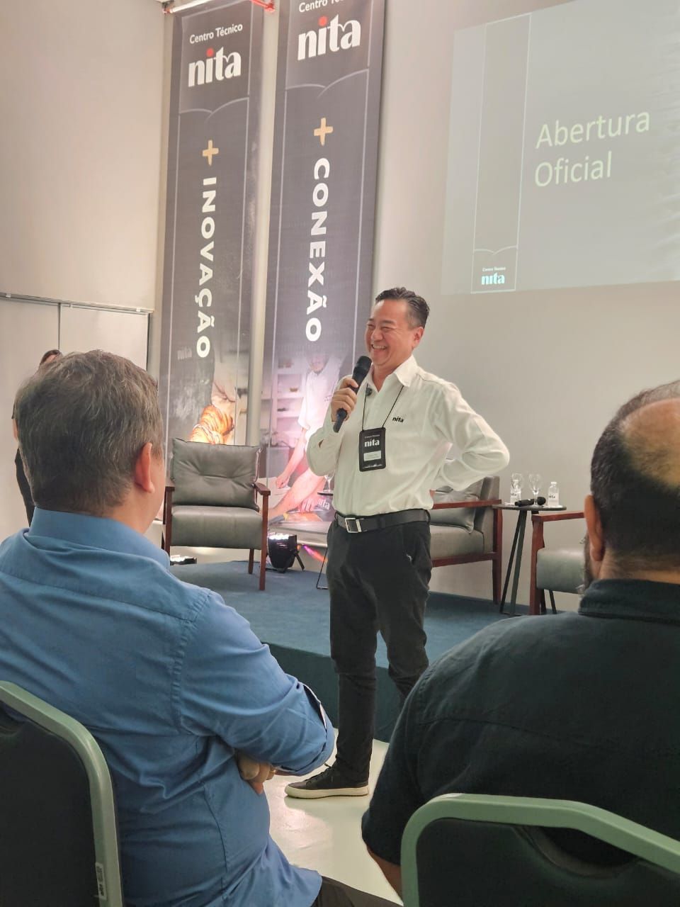 Homem fazendo um discurso em uma conferência, segurando um microfone, em pé na frente de uma plateia.