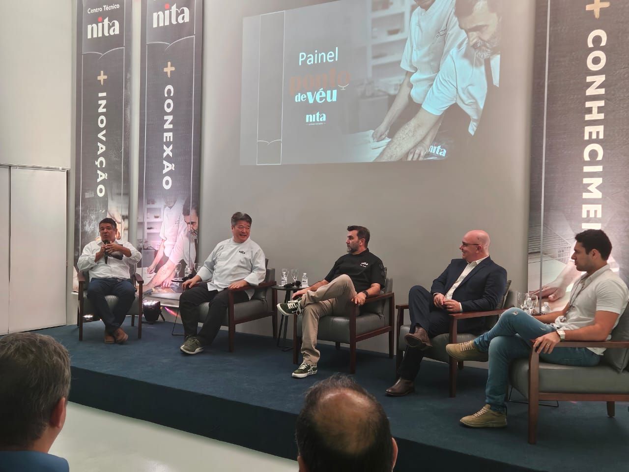 Painel de discussão em um evento com seis homens sentados no palco, discutindo inovação, conexão e conhecimento.