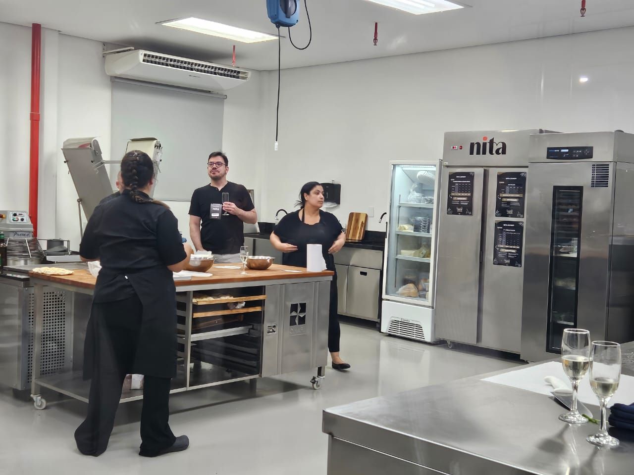 Cozinha de padaria: três pessoas trabalhando em um balcão com fornos e geladeiras.