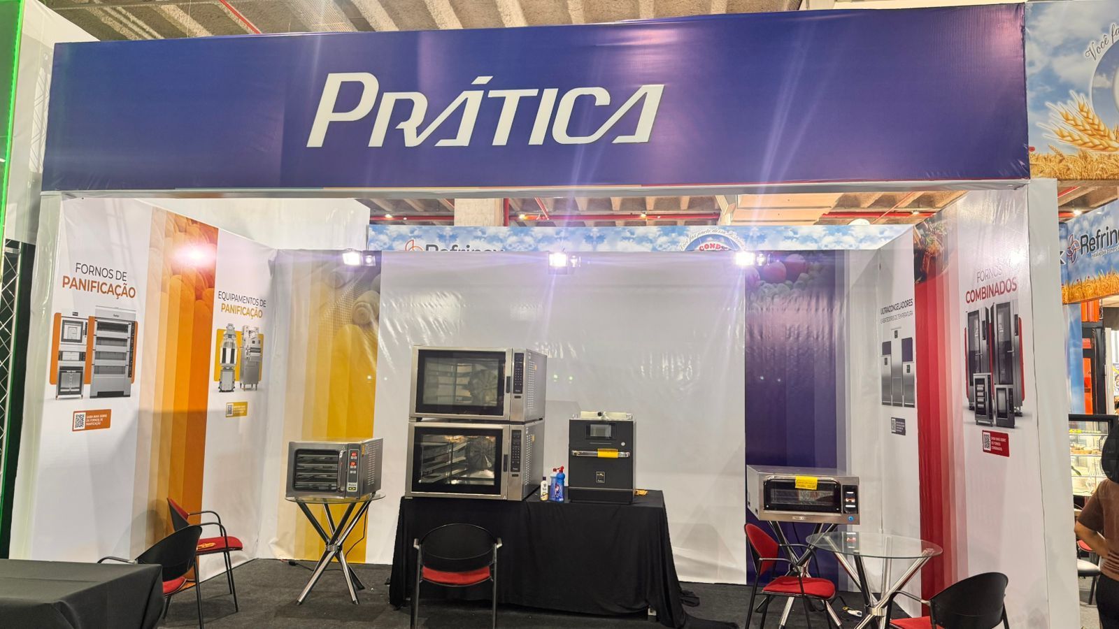 Estande da Prática: Placa azul, fornos comerciais em exposição, paredes transparentes, ambiente interno.