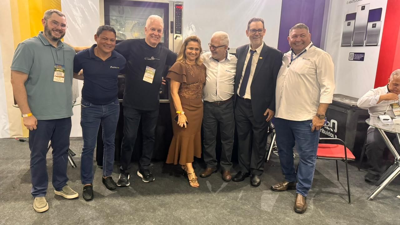 Grupo de pessoas juntas, sorrindo, em um estande de feira comercial, em ambiente fechado.