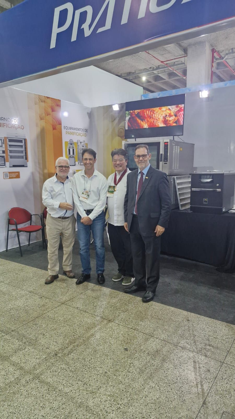Quatro homens estão em frente a um estande de uma feira, provavelmente em um evento do setor. Eles posam com um grande forno.