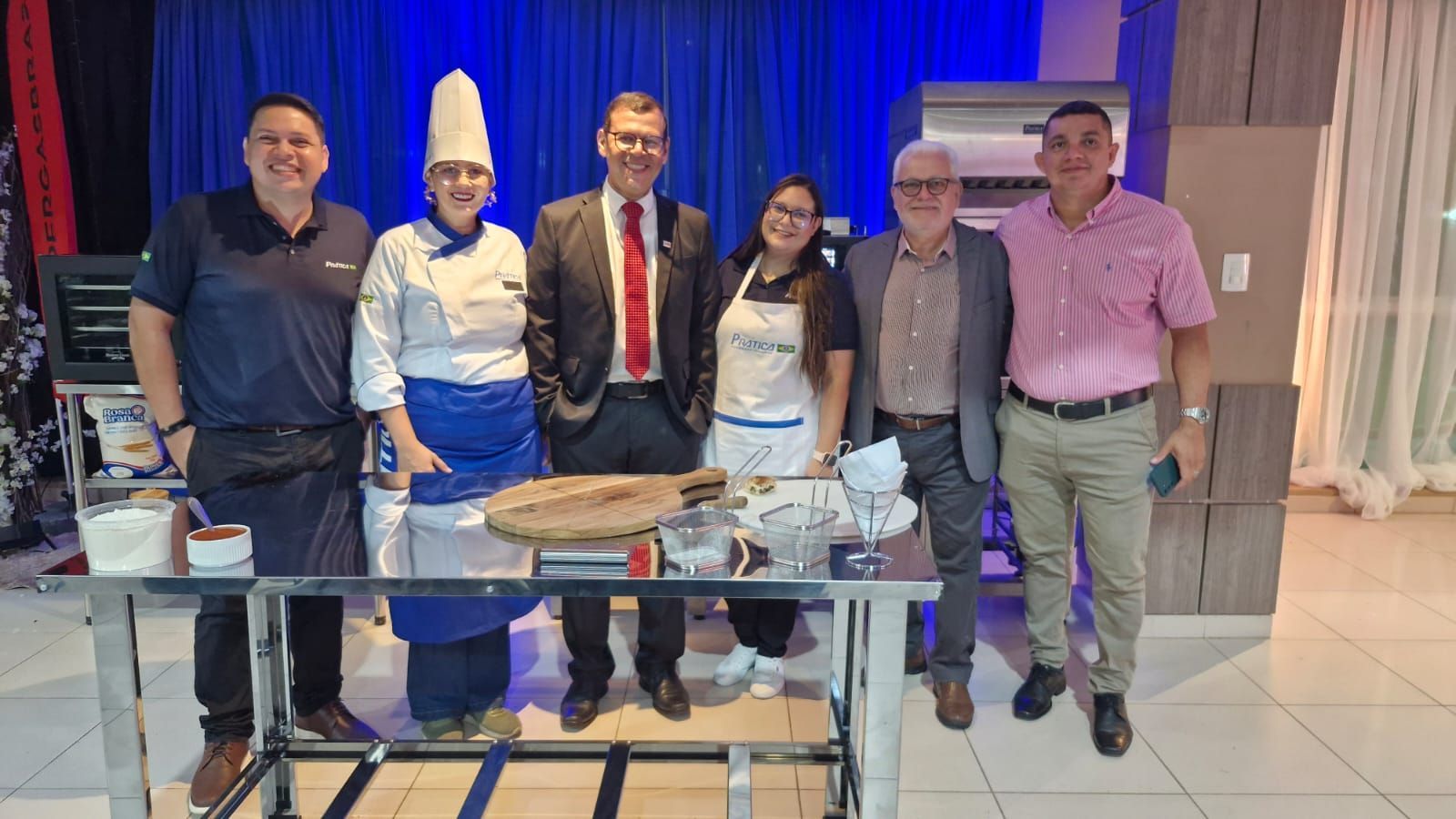 Grupo de pessoas em pé atrás de uma mesa com alimentos. Fundo azul, chapéu de chef, sorrisos.