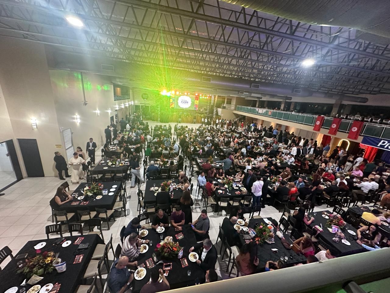 Grande evento em um salão com muitas pessoas sentadas em mesas, um palco iluminado com holofotes.