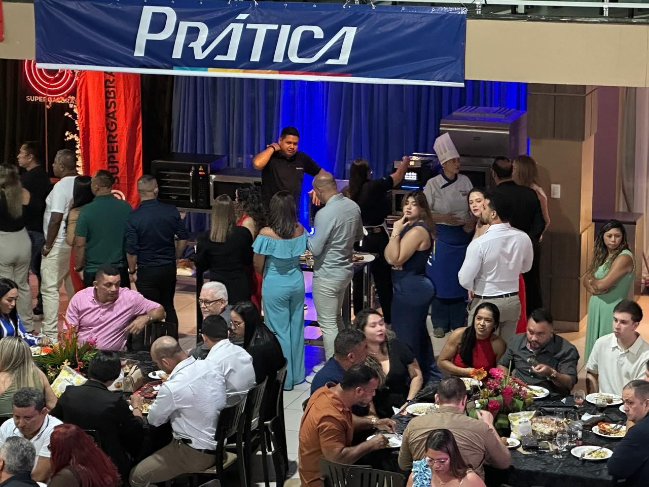 Pessoas em um evento com mesas, palco e faixa com os dizeres 