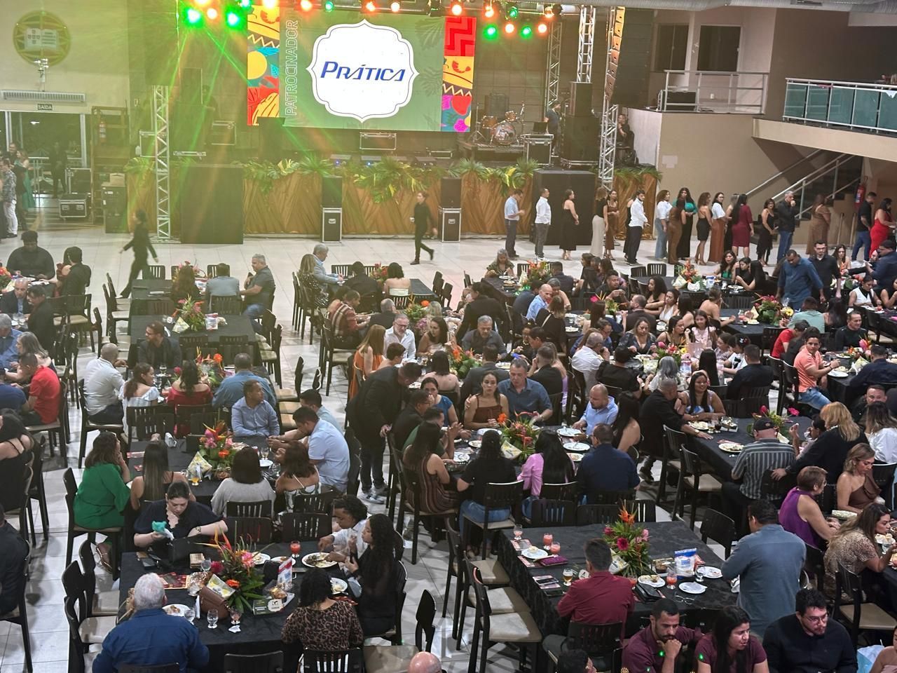 Grande evento interno com mesas, palco com banda e muitas pessoas reunidas.