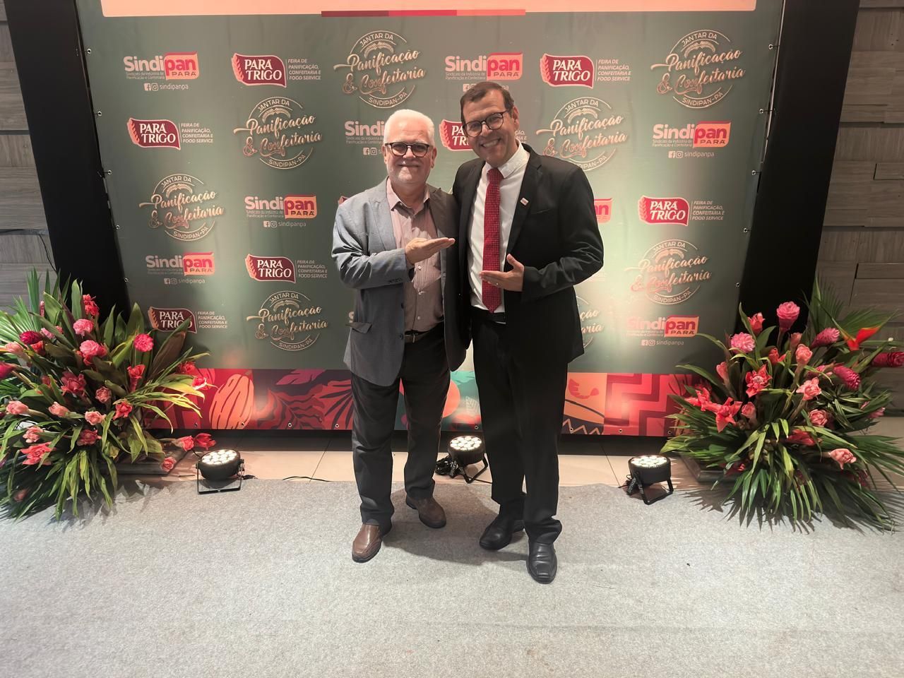 Dois homens posando em frente a um cenário com logotipos e flores. Um aponta. O outro sorri.