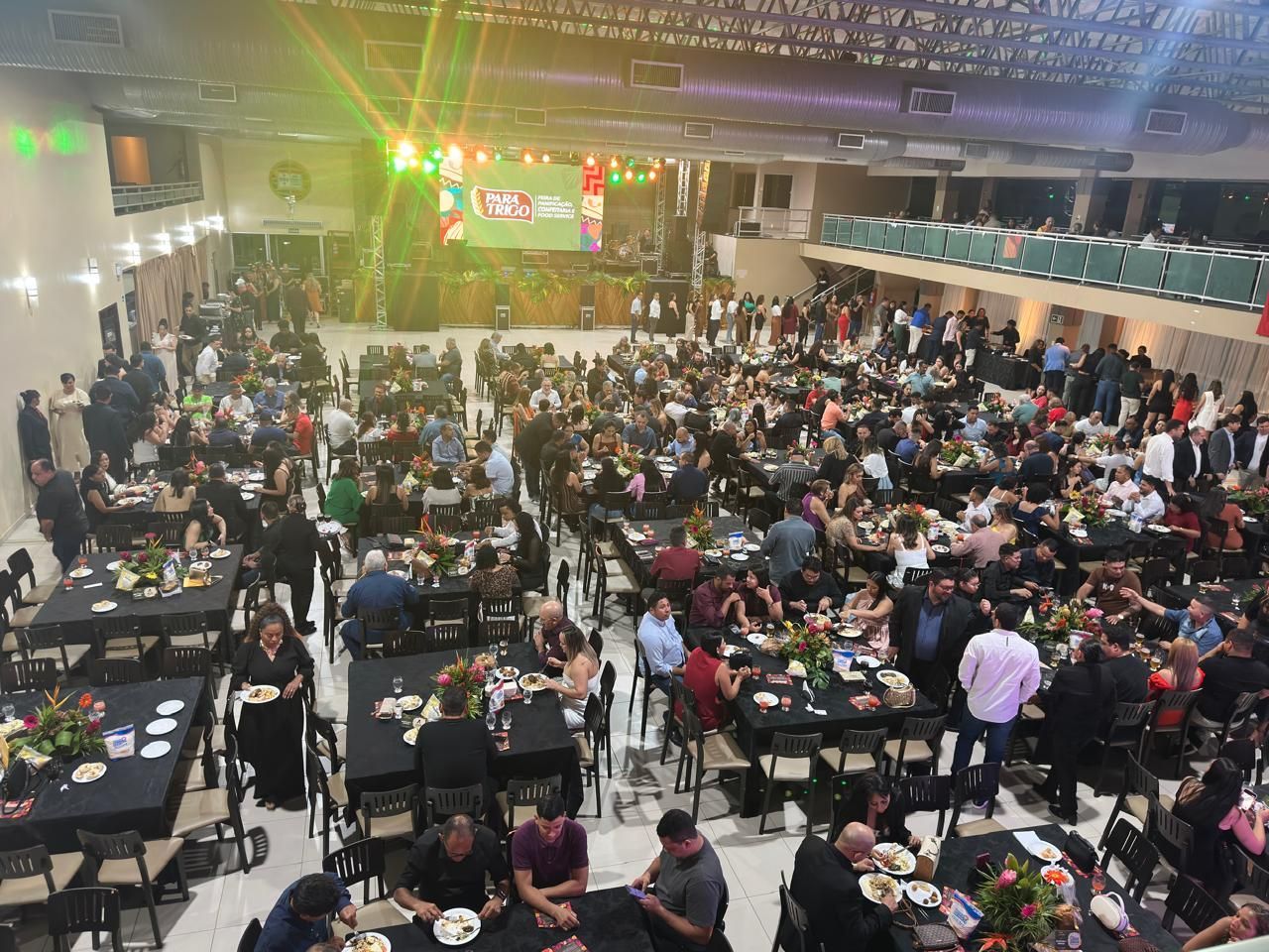 Grande salão de eventos com muitas pessoas sentadas em mesas e um palco ao fundo.