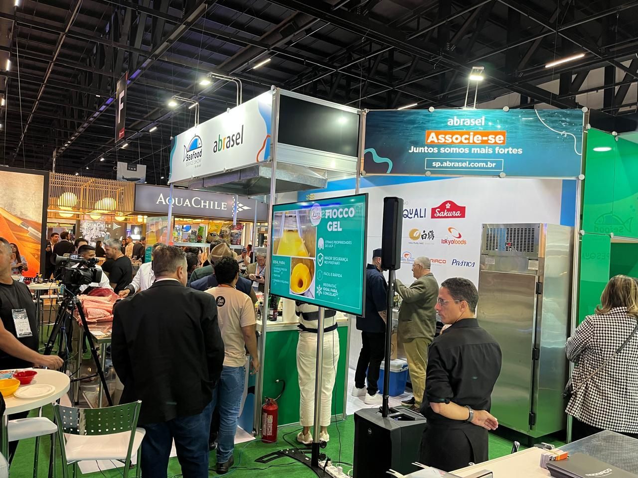 Pessoas em uma feira comercial com estandes e displays. Placas, telas e produtos visíveis.