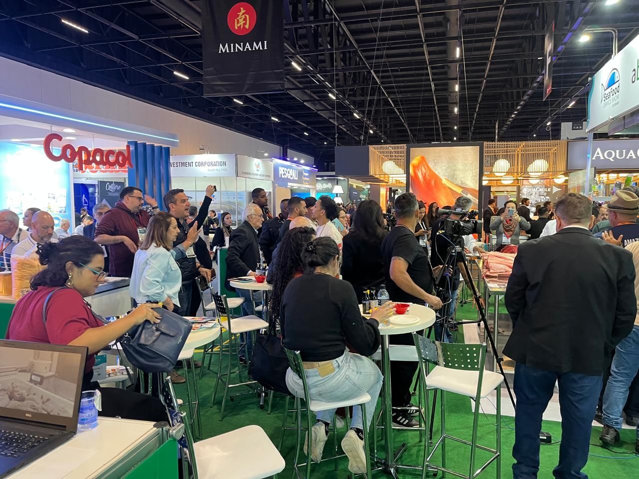 Feira com pessoas reunidas; algumas sentadas em mesas. Placas de sinalização do estande visíveis, luzes brilhantes.