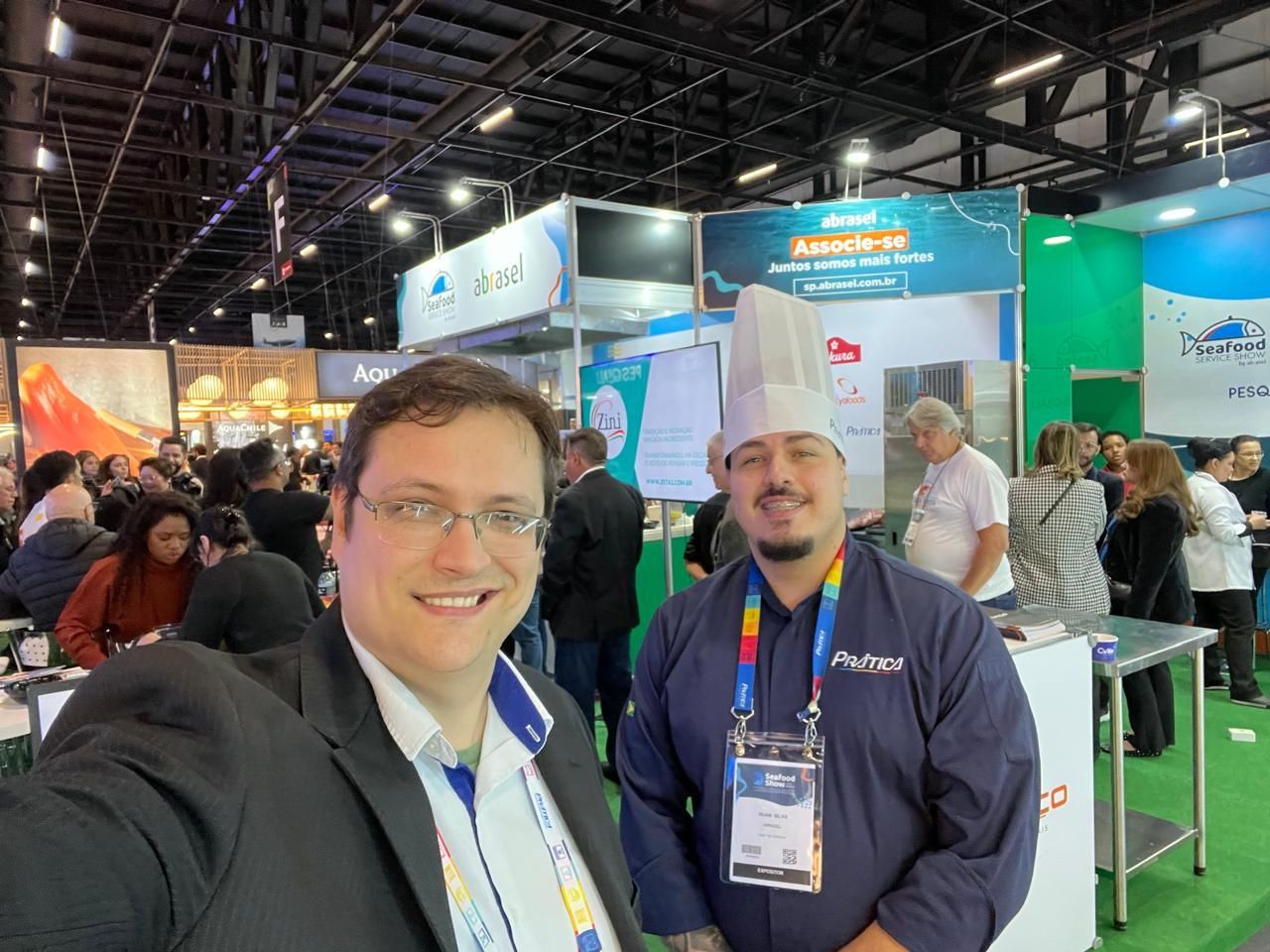 Dois homens sorrindo em uma convenção; um de terno, outro com chapéu e uniforme de chef. Eles estão em uma feira com muitos estandes.