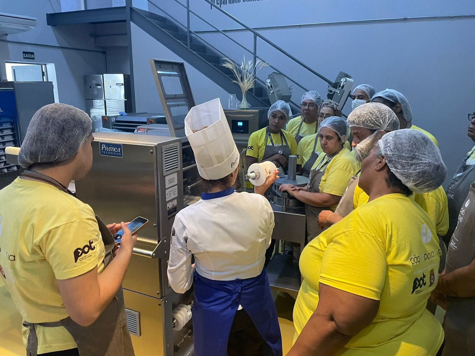 Chef demonstra equipamento para um grupo de pessoas usando toucas de cabelo e camisas amarelas em uma cozinha.