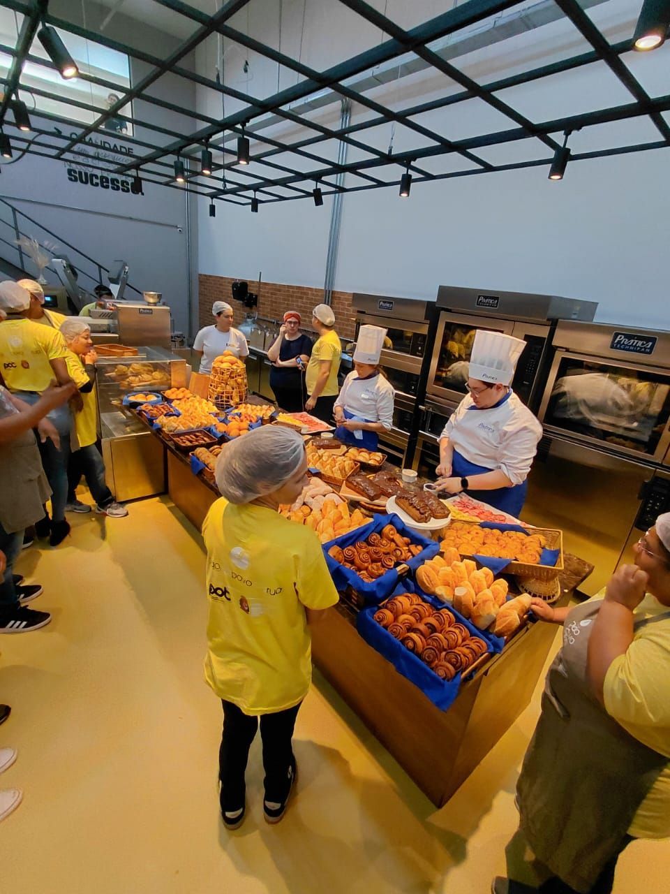 Exposição de padaria com vários doces e pessoas; funcionários usando chapéus de chef e camisas amarelas.