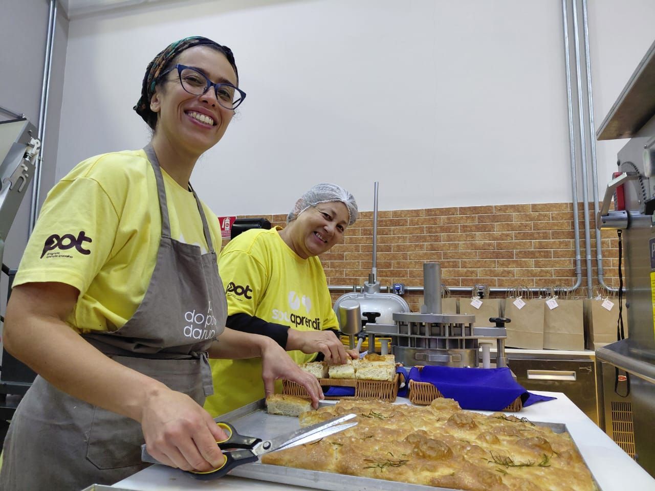 Duas pessoas de camisa amarela cortando pão na cozinha de uma padaria, sorrindo para a câmera.