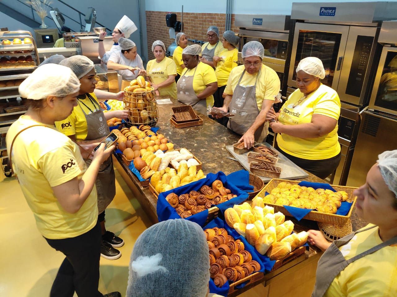 Pessoas com camisas amarelas e redes de cabelo ao redor de uma mesa de produtos assados em uma padaria.