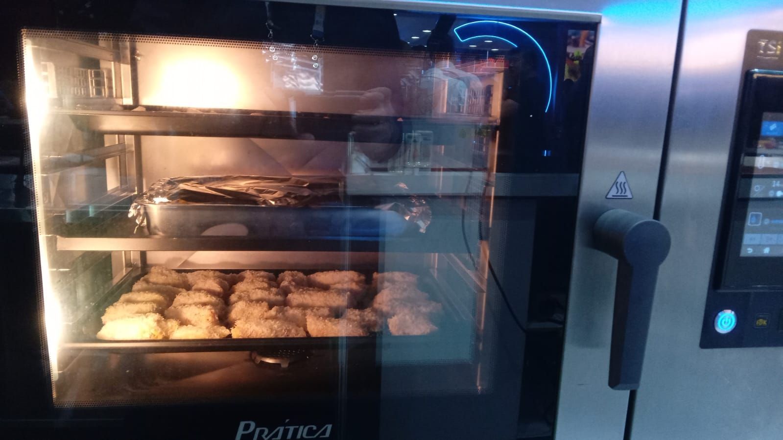 Forno comercial com alimentos dentro, incluindo bandejas de itens assados.