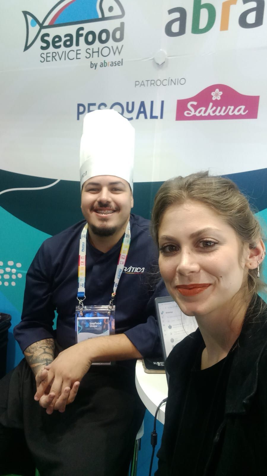 Chef e mulher sorrindo, em um estande da Seafood Service Show. Chef usa chapéu branco e uniforme azul.