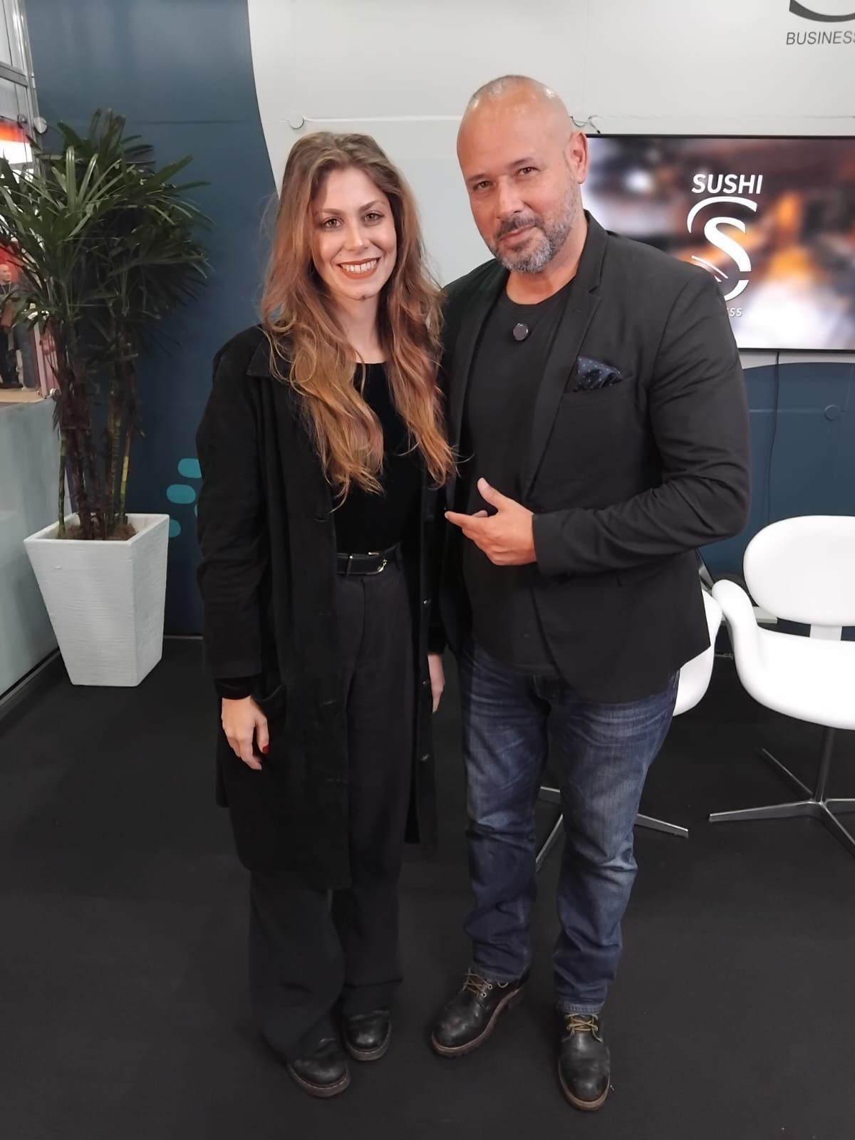 Mulher e homem posando. Mulher de roupa preta; homem de jaqueta preta e calça jeans. Ambiente interno.