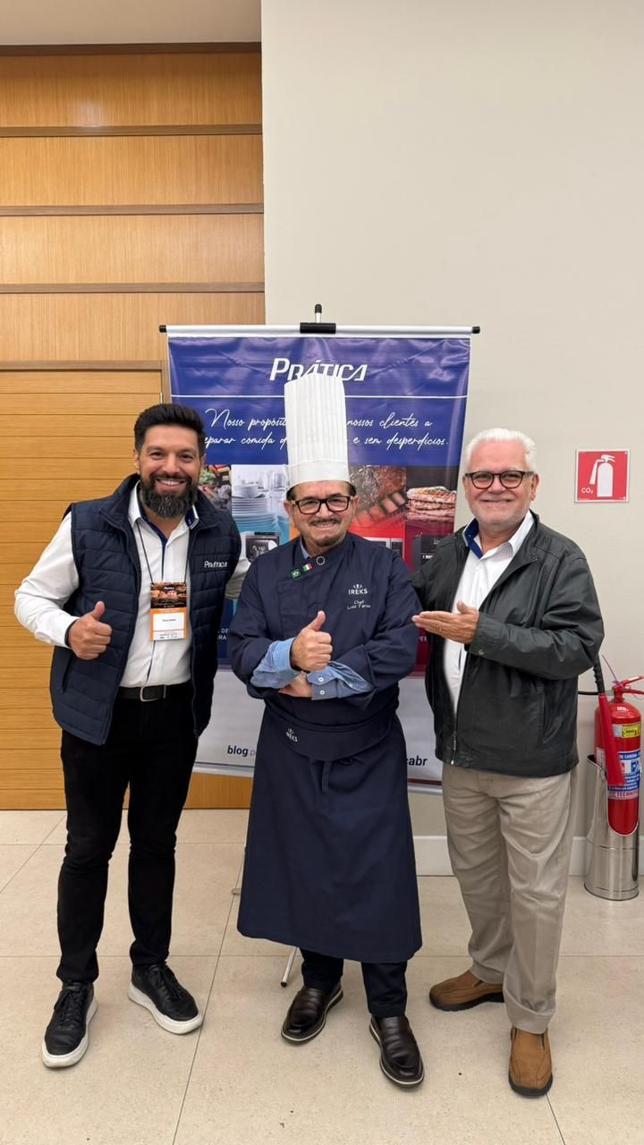 Três homens posam em frente a uma faixa; um deles, com uniforme de chef, faz sinal de positivo.