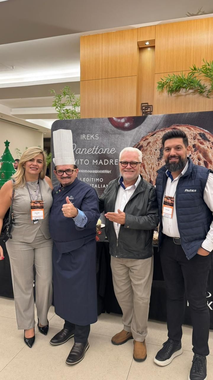 Quatro pessoas posando para foto em uma cabine. Uma delas, com uniforme de chef, faz sinal de positivo. Ao fundo, há um banner.
