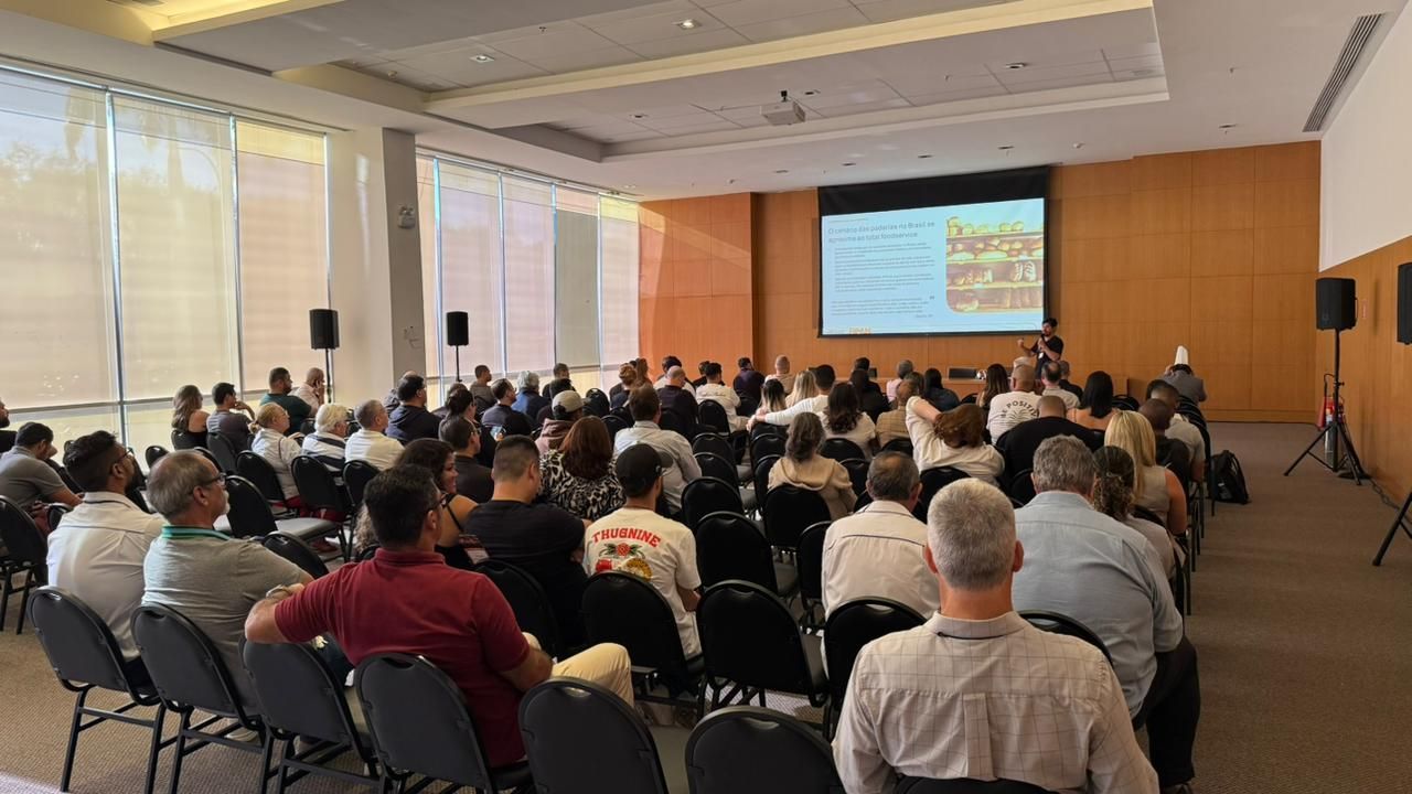 Público sentado em uma sala de conferências, de frente para uma tela com uma apresentação. Os palestrantes estão de cada lado.