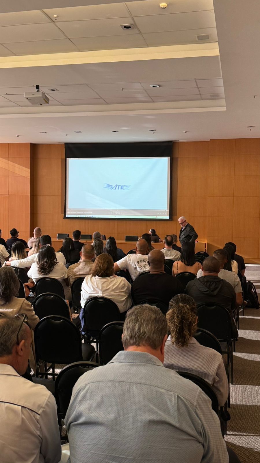 Uma apresentação em conferência: palestrante em um pódio, público sentado em fileiras, tela exibindo texto em uma sala grande.