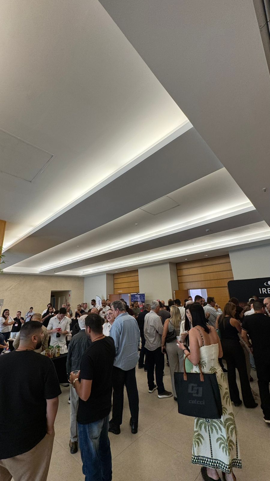 Multidão em uma sala bem iluminada com teto branco, em pé perto de mesas, algumas com displays.