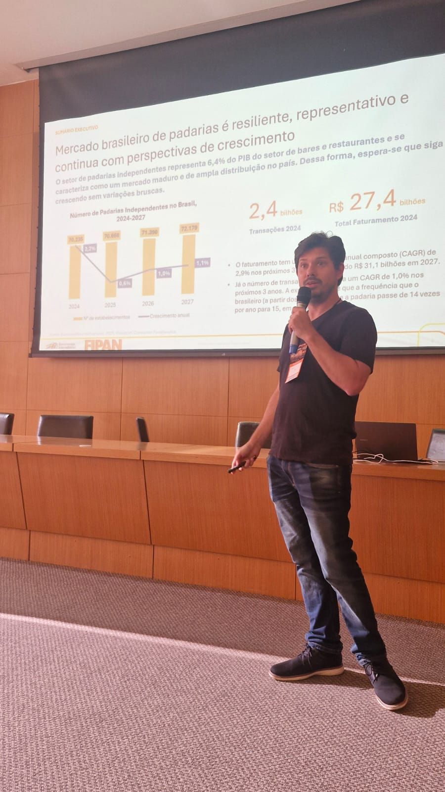 Homem apresentando-se em frente a uma tela, segurando um microfone. Ele veste calça jeans, camisa preta e tênis.