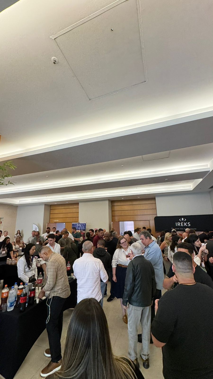 Sala grande e lotada; pessoas reunidas em volta de mesas, provavelmente em um evento.