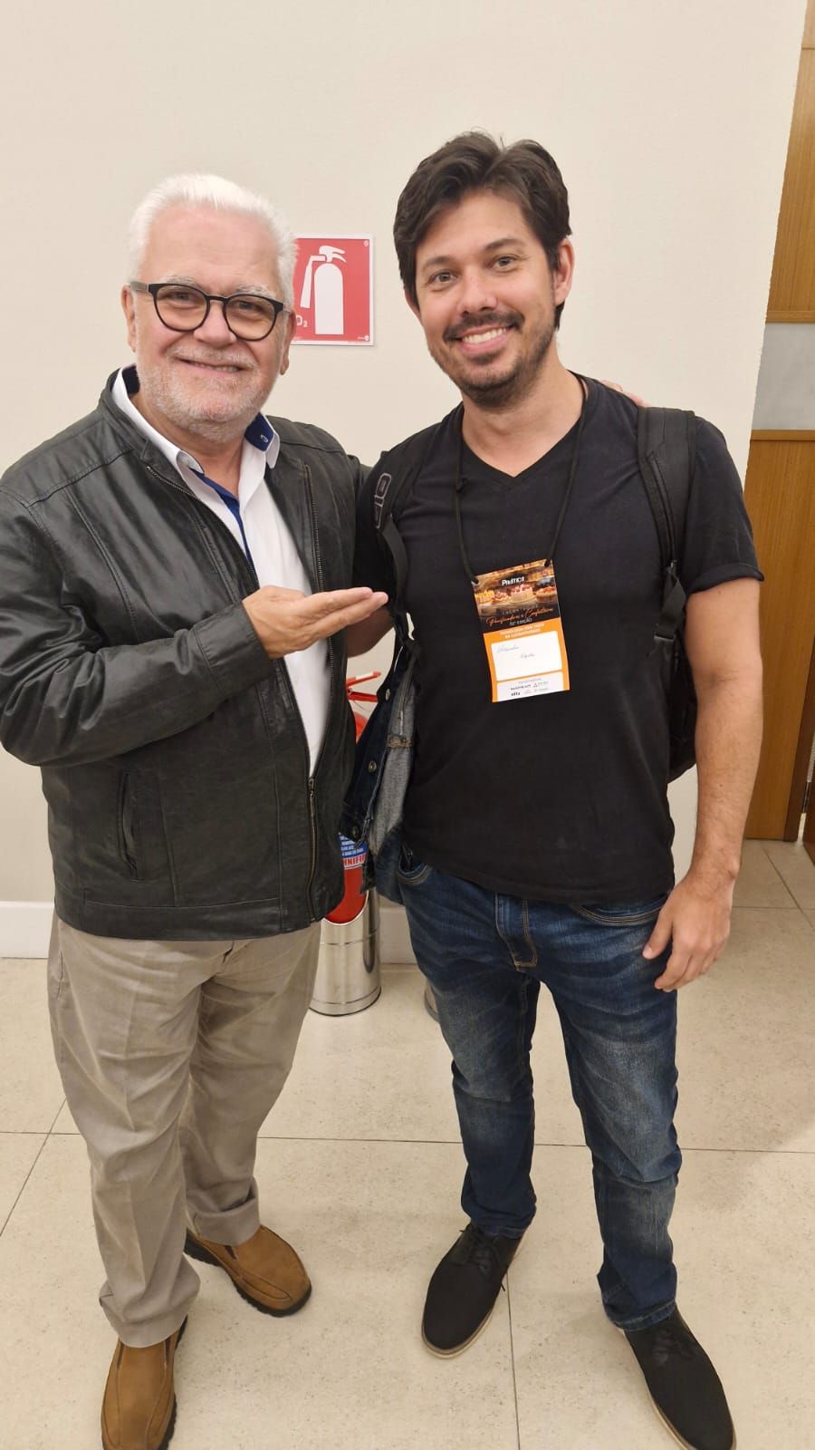 Dois homens juntos; um deles apontando e sorrindo. Interior, fundo neutro.