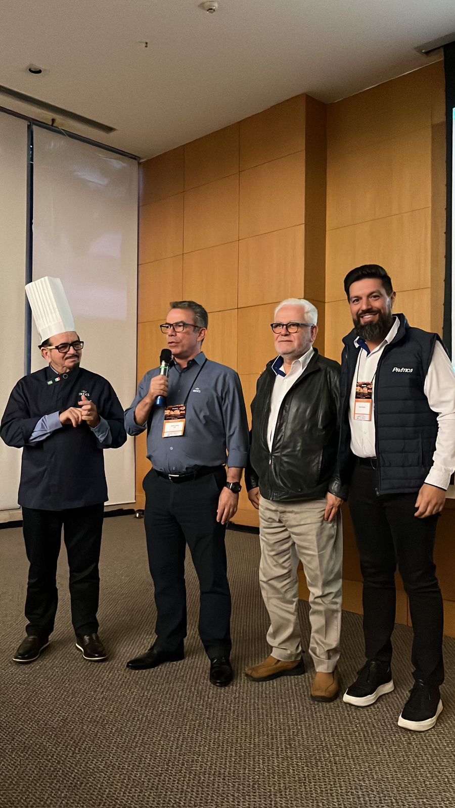 Quatro homens em uma sala; um com chapéu de chef, outros falando em um evento, um segurando um microfone.