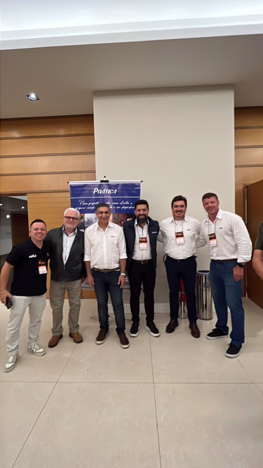 Grupo de sete homens em frente a uma faixa. Alguns usando crachás. Ambiente interno.