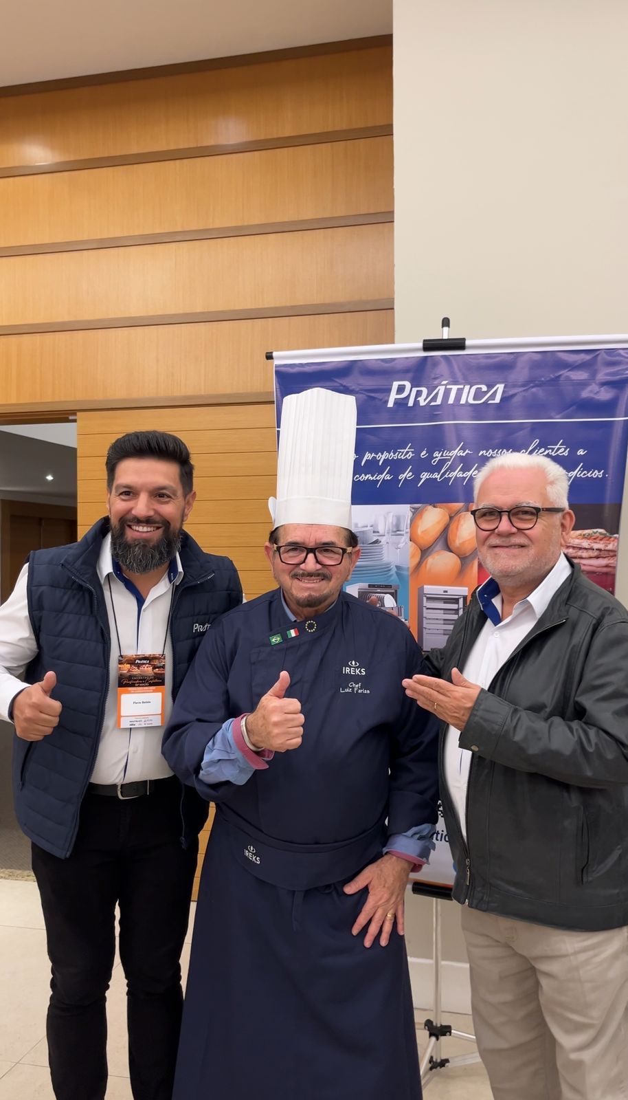 Três homens posam em frente a uma faixa. Um chef de chapéu branco e uniforme azul faz sinal de positivo. Os outros apontam.