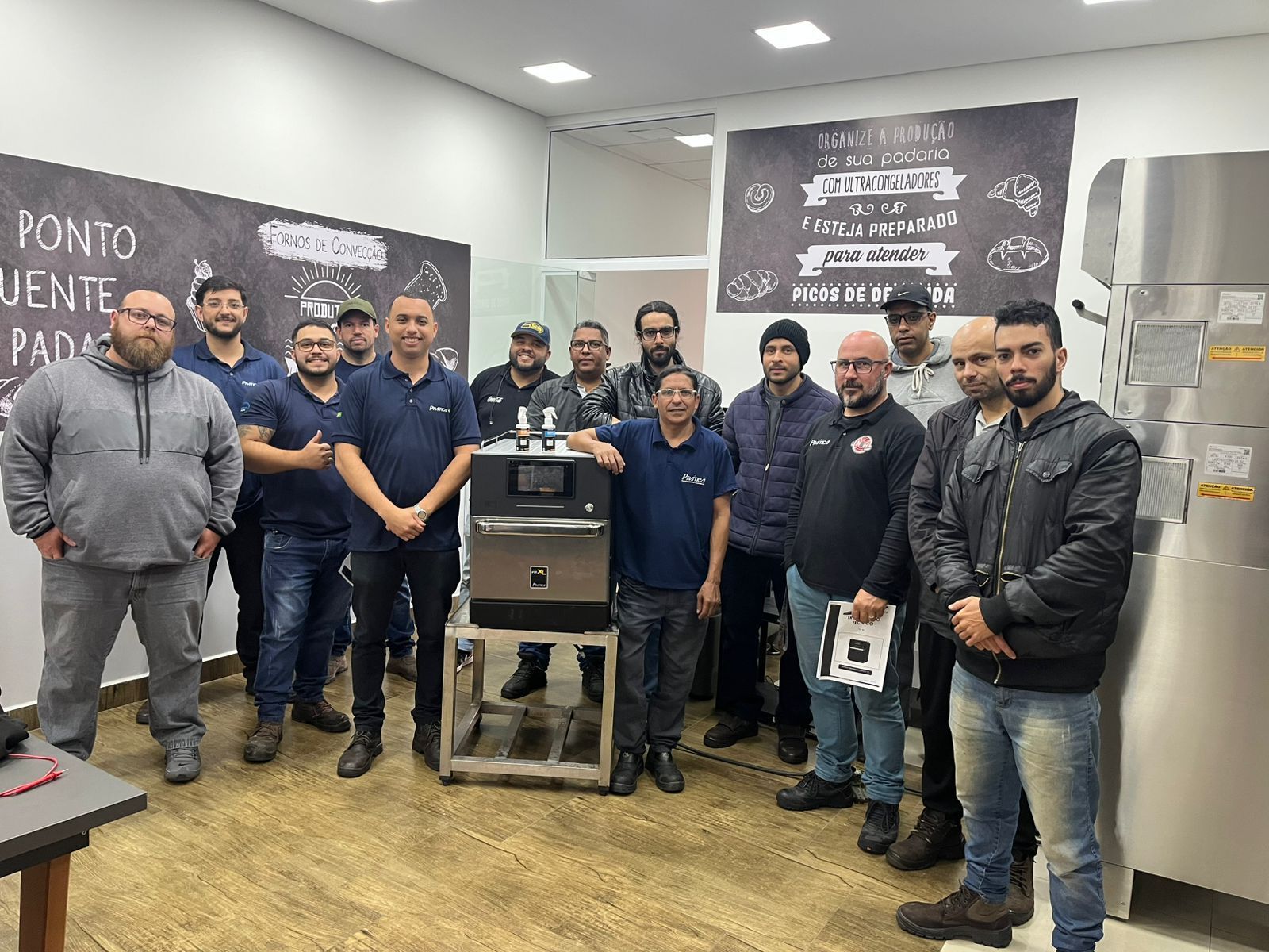 Grupo de homens em uma oficina, reunidos em volta de máquinas; alguns seguram equipamentos; faixas na parede.