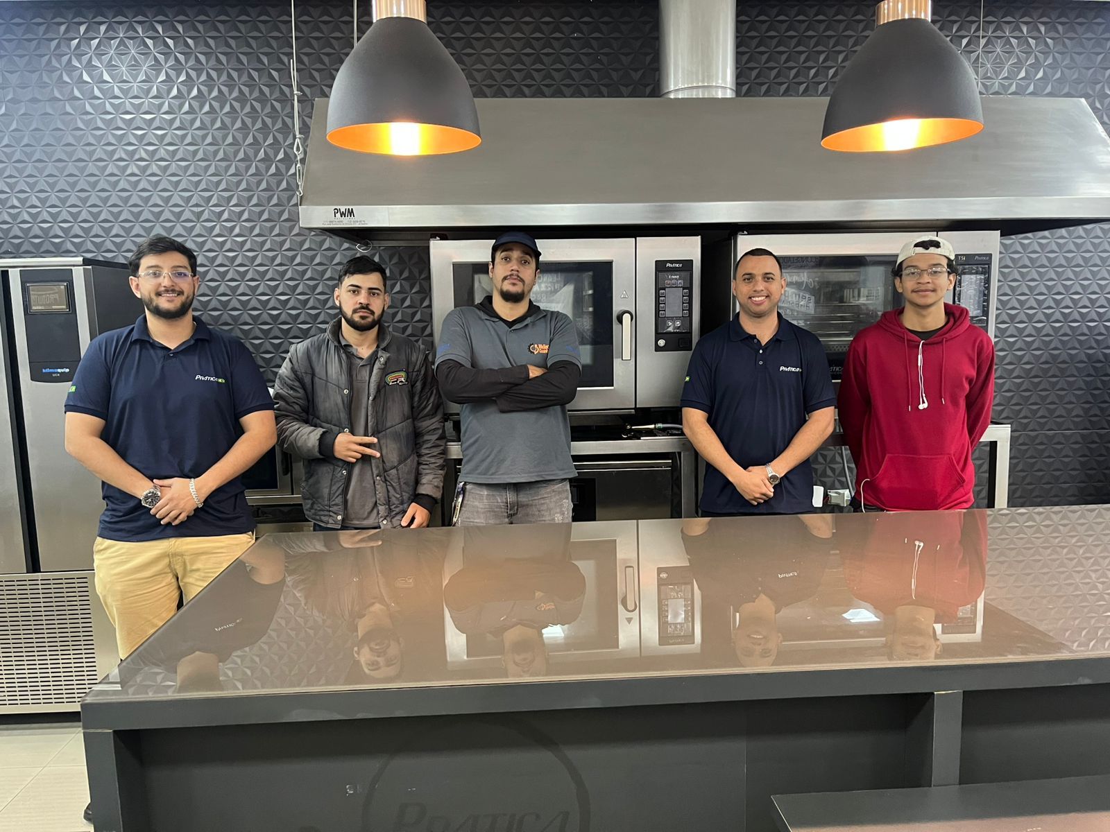 Cinco pessoas em pé em frente a equipamentos de cozinha. Dois homens em cada extremidade, outros no meio. Eletrodomésticos de aço inoxidável.